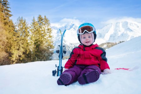 Photo for Little girl sit in snow with small mountain ski in sport outfit including helmet and skiing goggles over mountains - Royalty Free Image