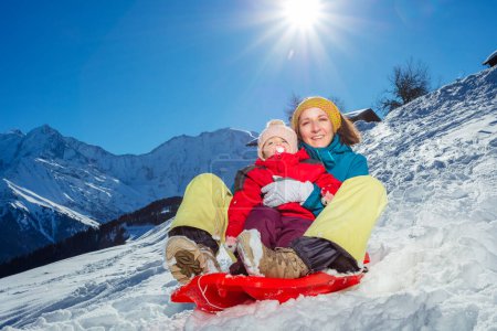 Téléchargez les photos : Mère avec bébé fille ensemble assis dans le traîneau descendre de la colline en hiver et de hautes montagnes sur le fond - en image libre de droit