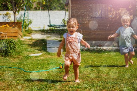 Foto de Diversión en el caluroso día de verano dos niños niño y niña corren alrededor del aspersor de agua en el césped en el jardín, saltando sobre chorros - Imagen libre de derechos