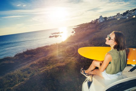 Téléchargez les photos : Vue d'en haut de la jeune femme assise sur le capot de la voiture regardant le coucher du soleil sur la mer - en image libre de droit