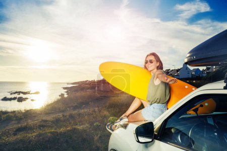 Foto de Young woman with surfboard sit on a car hood look over sea and sunset turn back to camera - Imagen libre de derechos