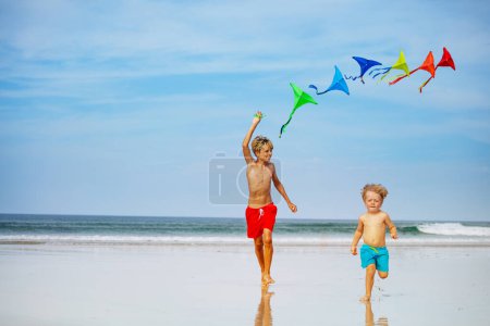 Foto de Dos niños felices, chicos corren sosteniendo muchas cometas coloridas en la playa sonriendo sobre el cielo durante las vacaciones - Imagen libre de derechos