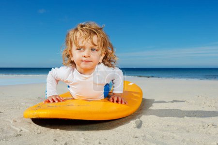 Téléchargez les photos : Belle petite fille blonde mignonne tout-petit pratiquant déjà de se lever et de pousser sur la planche de surf sur la plage - en image libre de droit