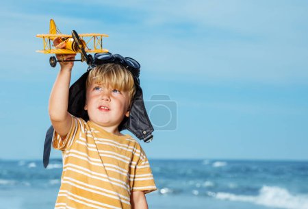 Cute smiling boy close portrait standing with toy model of the plane wearing aviation hat and googles on the beach