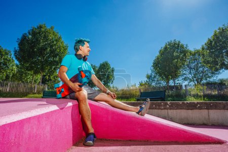 Téléchargez les photos : Man with blue hair sit on the pink ramp at the skatepark holding skate board on sunny summer day - en image libre de droit