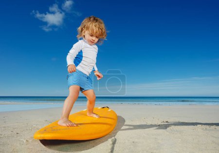 Foto de Little blond toddler girl already practicing and posing balancing standing on the surfboard on the beach - Imagen libre de derechos