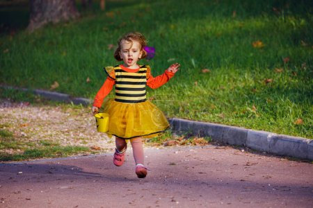 Foto de Happy little girl in Halloween bee costume disguise run on the road holding small bucket for candies - Imagen libre de derechos
