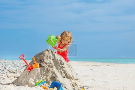 Téléchargez les photos : Happy little beautiful girl play with sand pouring water from plastic bucket on the sea beach during vacation - en image libre de droit