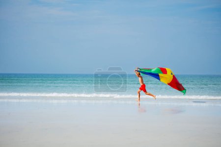 Téléchargez les photos : Jeune garçon avec tissu de parachute coloré volant sur le vent courir sur la plage mouvement dynamique action concept vue de profil latérale - en image libre de droit