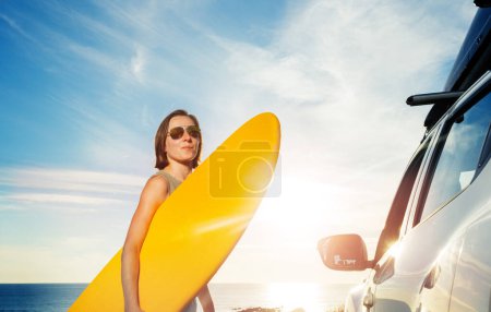 Téléchargez les photos : Jeune femme en lunettes de soleil marche avec planche de surf à la voiture sur le ciel du coucher du soleil - en image libre de droit
