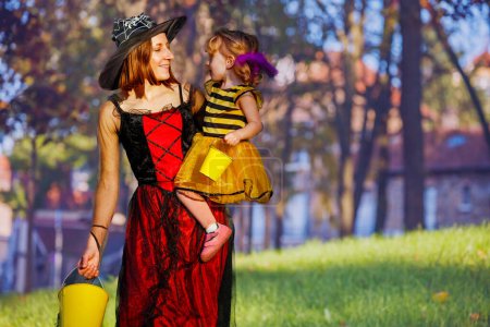 Téléchargez les photos : Portrait de mère tenant petite belle fille en costumes de sorcière et d'abeille Halloween avec des seaux pour tromper ou traiter des bonbons - en image libre de droit