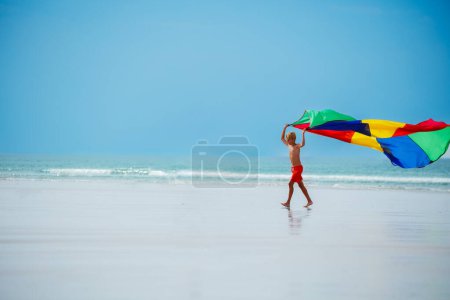 Téléchargez les photos : Joyeux garçon courir tenant tissu parachute coloré volant sur le vent sur la plage océan vue de profil latérale - en image libre de droit