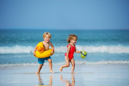 Foto de Cute little boy with girl run holding toy inflatable buoys duck and plastic bucket having fun running on the sand beach - Imagen libre de derechos
