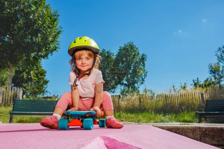 Foto de Little beautiful girl sit on the skate playing at skatepark wearing helmet on sunny summer day - Imagen libre de derechos