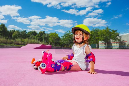 Téléchargez les photos : Petite fille mignonne de deux ans apprendre à patiner sur rouleaux, assis avec casque et protection sur le plancher de couleur du parc - en image libre de droit