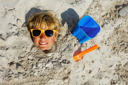 Photo for Boy's face dug into sand at the beach with all covered but the head wearing sunglasses, smile, toy bucket and scoop aside top view - Royalty Free Image