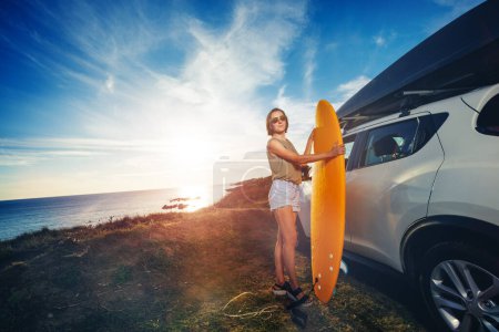Portrait d'une jeune femme en lunettes de soleil se tenir avec planche de surf par la voiture au coucher du soleil regarder à la caméra