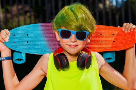 Téléchargez les photos : Calme portrait en gros plan tendance d'un garçon avec skateboard sur les épaules écouteurs de cheveux verts et portant des lunettes de soleil - en image libre de droit