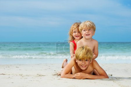 Foto de Group of three happy boys and a girl children lay on top of another on the sea sand beach at summer vacation - Imagen libre de derechos