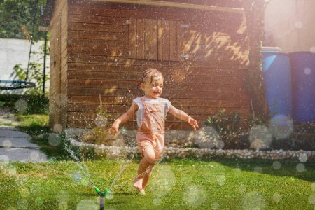 Téléchargez les photos : Joyeux belle fille en vêtements humides courir autour avec arroseur d'eau dans la pelouse du jardin sur la chaude journée d'été - en image libre de droit