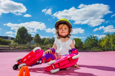Téléchargez les photos : Smiling little one year old girl learn to skate on rollers, sit with helmet and protection on the color floor of the park - en image libre de droit
