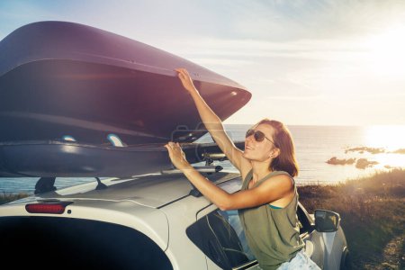 Téléchargez les photos : Portrait of a young woman in sunglasses unload car roof top baggage rack standing on the vacations parking over ocean sunset - en image libre de droit