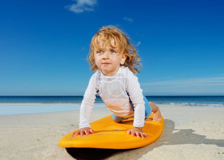Photo for Little blond toddler girl already practicing to stand up and push up on the surfboard on the beach - Royalty Free Image