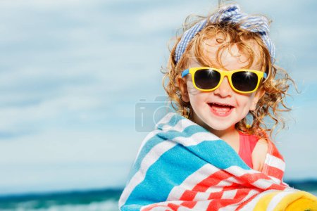 Téléchargez les photos : Portrait serré d'une petite belle fille assise enveloppée dans une serviette de plage sur un caillou à la mer souriant et regardant la caméra - en image libre de droit