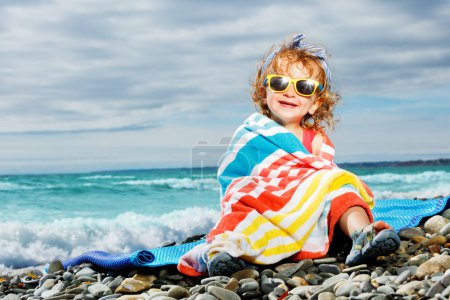 Téléchargez les photos : Little beautiful girl sit wrapped in beach towel on pebble at the ocean smiling and looking at camera - en image libre de droit