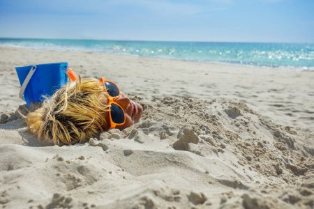 Photo for Boy's face dug into sand on the beach with all covered but the head wearing sunglasses, smile, toy bucket and scoop aside over sea - Royalty Free Image