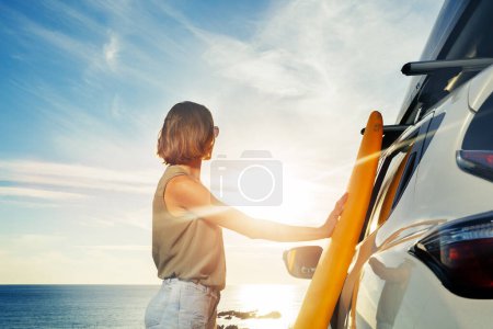 Foto de Young woman stand with surf board by the car looking over ocean sunset view from behind - Imagen libre de derechos