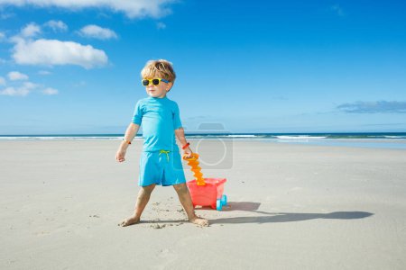 Foto de Niño feliz en gafas de sol caminar en la playa de arena blanca jugando con carro de juguete - Imagen libre de derechos