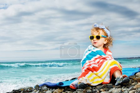 Téléchargez les photos : Petite belle fille assise enveloppée dans une serviette de plage sur un caillou à la mer dans des lunettes de soleil - en image libre de droit