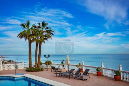 Téléchargez les photos : Ambiance de vacances avec chaises longues près de la piscine et des palmiers sur la mer Méditerranée calme - en image libre de droit