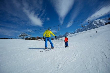 Photo for Adult on skiing slope glide downhill teaching little child to ski both holding poles - Royalty Free Image