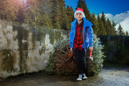 Téléchargez les photos : Joyeux jeune garçon avec hache à la main dans le chapeau de Père Noël ramener à la maison arbre de Noël pour la célébration du Nouvel An tenant tronc et souriant - en image libre de droit