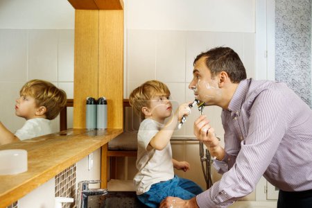 Photo for Little kid sitting on sink in sunny bathroom holding razor and helping his father to shave before work - Royalty Free Image