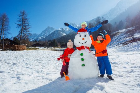 Photo for Happy kids, boys and a girl construct snowman play outside in the mountainous region posing lifting hands - Royalty Free Image