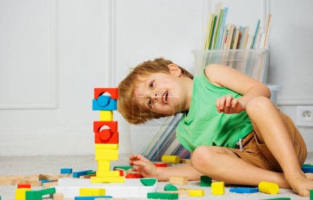 Foto de Riéndose Niño sentado en una alfombra en una sala de estar, mira su torre de juguete de bloques de madera, rodeado de cajas de juguetes y libros - Imagen libre de derechos