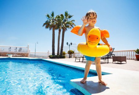 Photo for Cute kid with floaties and yellow inflatable duck ring eager to jump in the pool waving hand over blue sky - Royalty Free Image