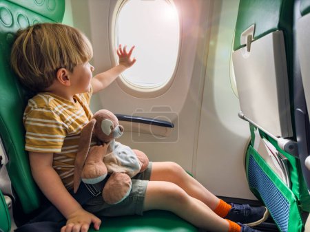 Foto de Guapo niño se sienta cerca de la ventana en el avión abrazo peluche juguete sosteniendo ojo de buey con la mano - Imagen libre de derechos