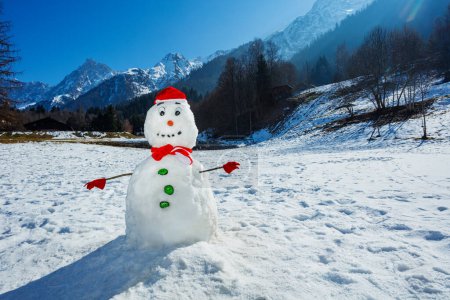 Photo for In a winter field surrounded by mountains,snowman in a scarf Santa hat red mittens a carrot nose and a charming smile, gazes out with stunning eyes - Royalty Free Image