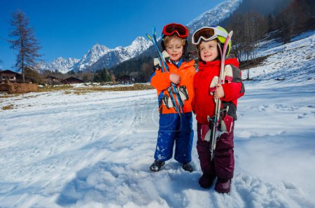 Foto de Dos niños niños y niñas en su primera escapada alpina, usan cascos, máscaras de esquí mientras están de pie con esquís en el terreno de montaña cubierto de nieve - Imagen libre de derechos