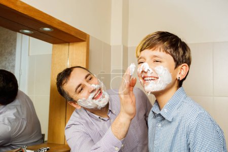 Téléchargez les photos : Père mousse visage de l'adolescent avec sa main se préparant pour le premier rasage, souriant ensemble dans la salle de bain - en image libre de droit