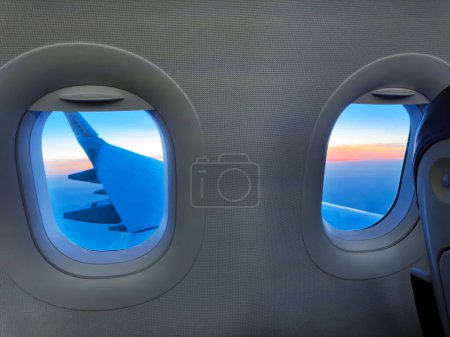 Téléchargez les photos : Vue de la fenêtre hublot dans un avion avec aile à l'extérieur et beau coucher de soleil à haute altitude - en image libre de droit