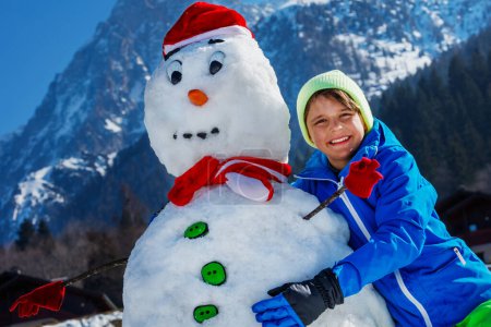 Téléchargez les photos : Gros plan portrait d'un jeune garçon heureux en tenue de sport d'hiver jouer avec bonhomme de neige dans le chapeau de Père Noël à l'extérieur - en image libre de droit