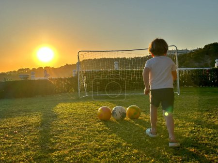 Photo for Little boy play football on lawn in the garden in sunset lights view from behind - Royalty Free Image