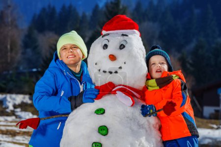 Photo for Close-up portrait of two happy boys brothers in winter sport outfit play with snowman in Santa hat outside - Royalty Free Image