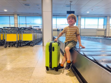 Photo for Handsome small boy sit and wait with hand suitcase on the baggage conveyor belt waiting for checked in luggage - Royalty Free Image
