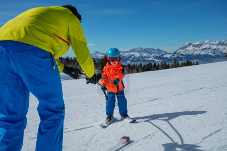 Photo for Dad teach little boy child to ski on the alpine slope by rolling below and holding poles - Royalty Free Image